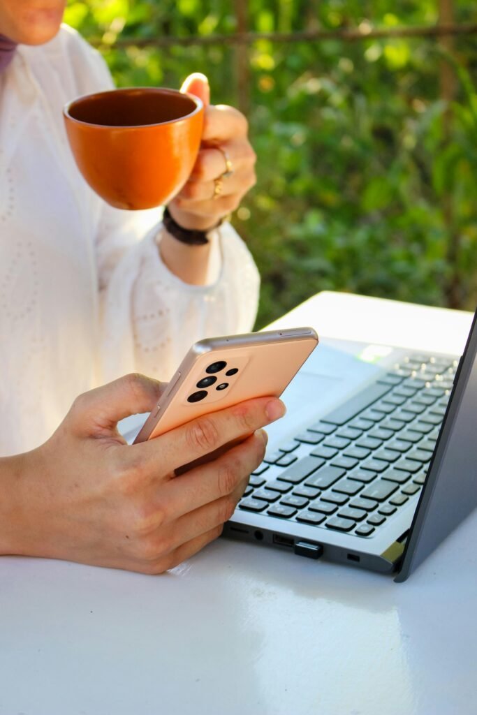 pexels-photo-17781733-17781733 Person using a smartphone while drinking coffee near a laptop outdoors, showcasing multitasking with technology.